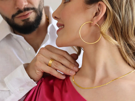  Un hombre usa en su dedo anular un anillo y acomoda una gargantilla tipo espejo en el cuello de una mujer, la cual también lleva puesto una candonga en oro amarillo de 18 quilates en su oreja.