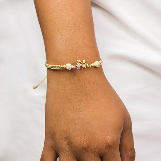 Mano de mujer con pulsera tejida en hilo beige coreano y accesorios de bisutería con el mismo color del hilo y dije con forma de niño, niña y perrito en oro amarillo de 18 quilates con cristales de color blanco, hecho en Joyería Caracas.