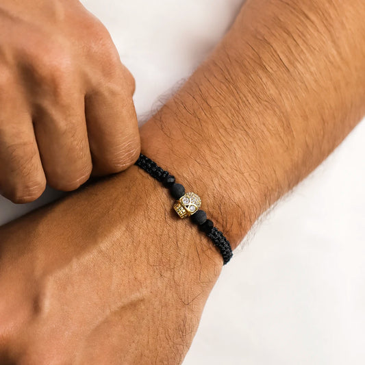 Mano de hombre con pulsera tejida en hilo negro terlenca, accesorios en bisutería de color negro y dije en forma de calavera en oro amarillo de 18 quilates con cristales de color blanco, hecho en Joyería Caracas. 