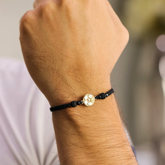 Mano de hombre con pulsera tejida en hilo negro terlenca, accesorios en bisutería de color negro y dije en forma de balón de fútbol con el numero cinco en el centro en oro amarillo de 18 quilates, hecho en Joyería Caracas. 